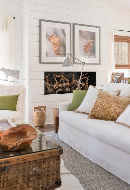 Białe panele z drewna na ścianie z kominkiem,dekoracje z korzeni,biala sofa z beżowymi i zielonymi poduszkami,wiklinowa skrzynia ze szklanym blatem w roli stolika w salonie
