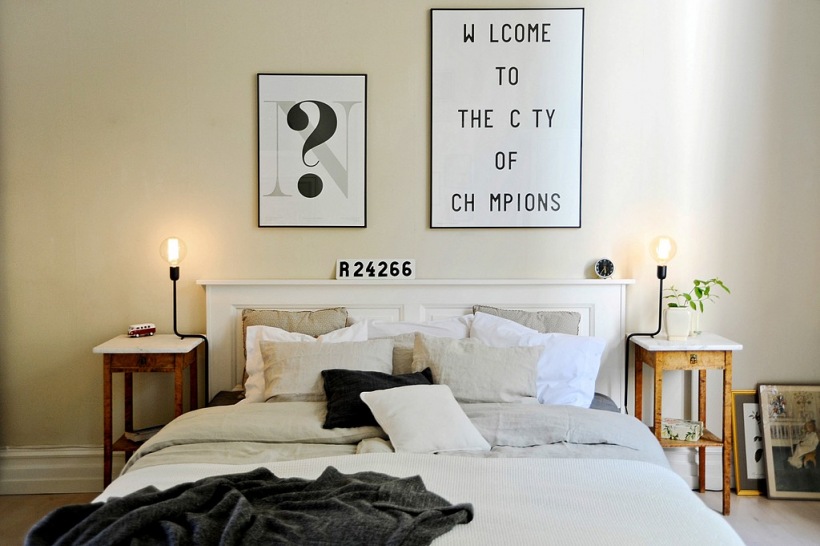 Typografie i grafiki na ścianie w skandynawskiej sypialni z białym łóżkiem i stylowymi drewnianymi stolikami nocnymi