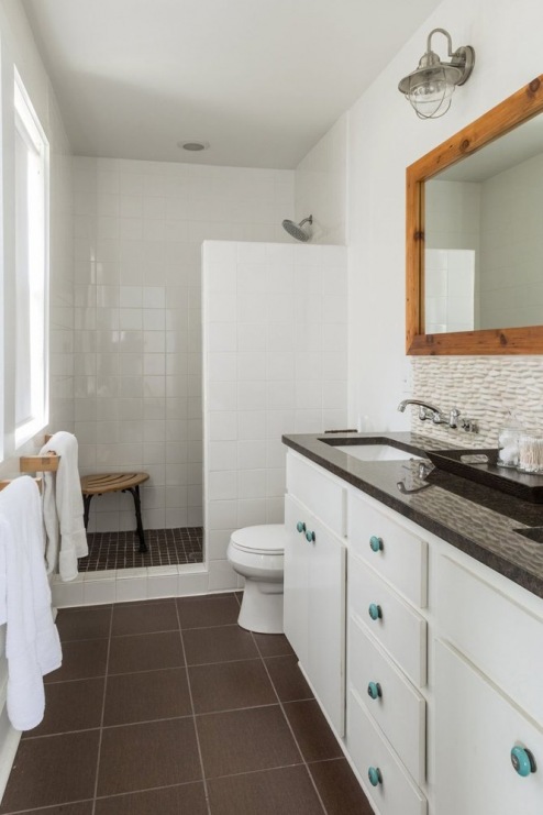 Higieniczną biel łazienki ociepla drewniana rama lustra i brązowa podloga oraz blat. Jasnozielone gałki szafek i...