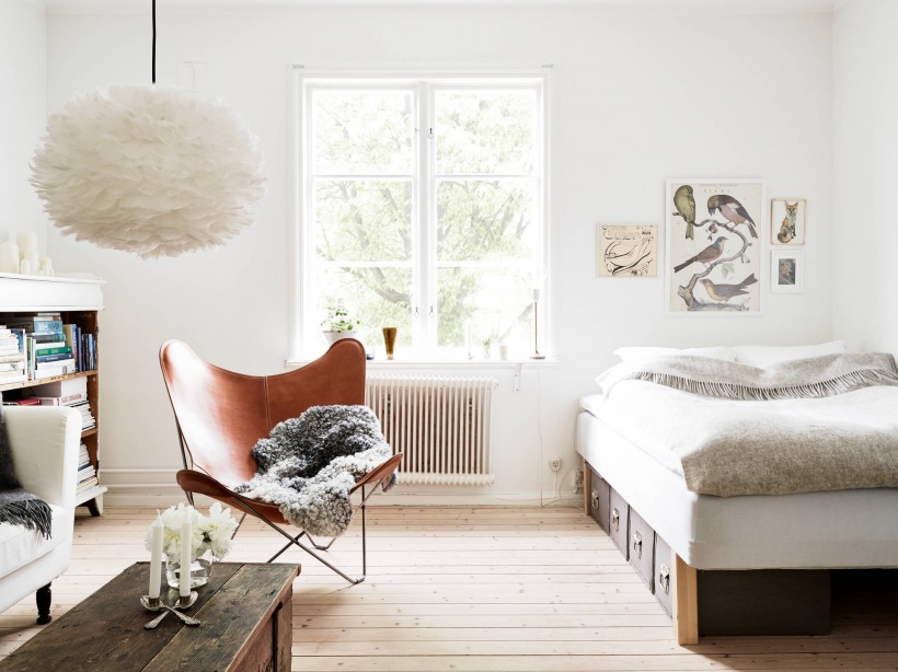 Lampa pompon,drewniana skrzynia,fotel skórzany motyl i łóżko w salonie małego mieszkania
