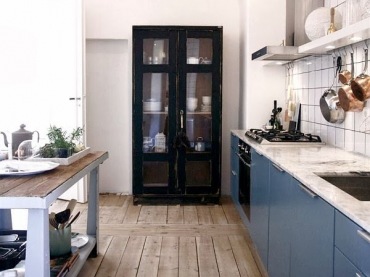 Niebieskie fronty szafek i drewniana podłoga  w kuchni z  czarną szafą tworzą wyśmienitą aranżację.Blat wyspy nawiązuje...