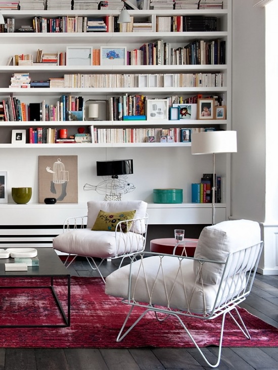 Różowy dywan vintage,biały nowoczesny fotel z metalowych pretów i z poduchami,białe półki na ścianie w salonie