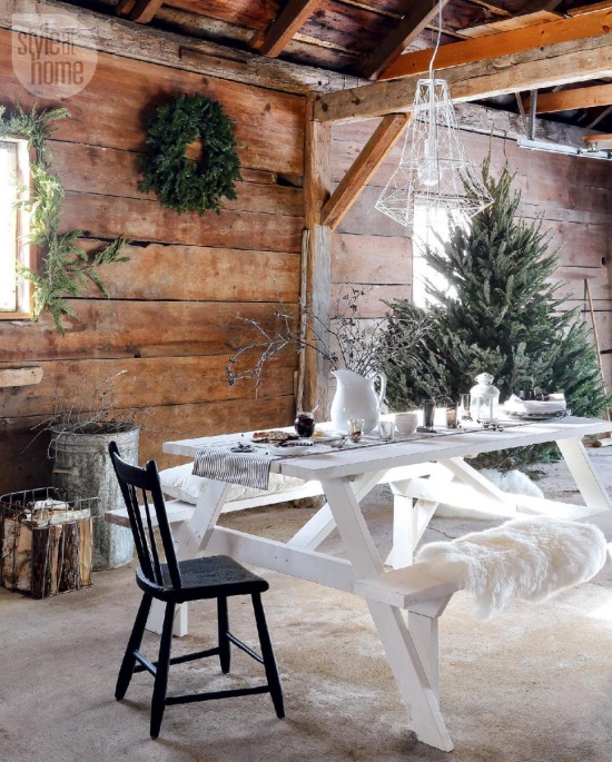 Białe druciane lampy nad malowanym białym stołem na kozłach z ławką,zielone girlandy i wianki z igliwia w aranzacji zimowego świątecznego tarasu