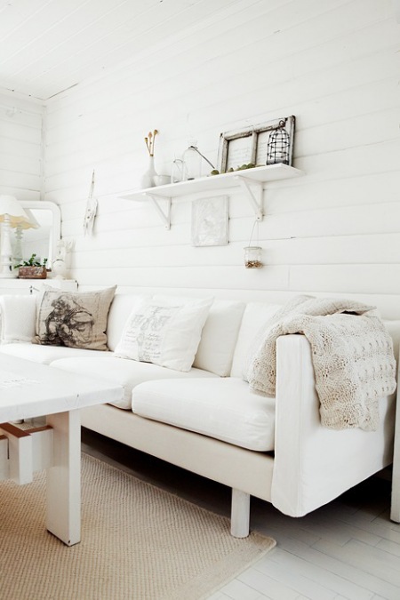 Skandynawska biała kanapa, białe drewniane pólki na scianie i biały drewniany stół w wiejskim salonie