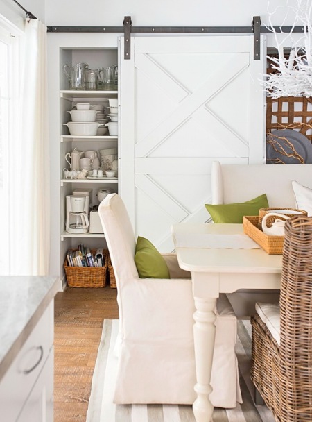Białe drzwi wrota na żelaznych szynach przy białym regale z półkami w aranżacji jadalni z białym stołem,krzesłami w sukienkach i wiklinowym krzesłem z poduchą