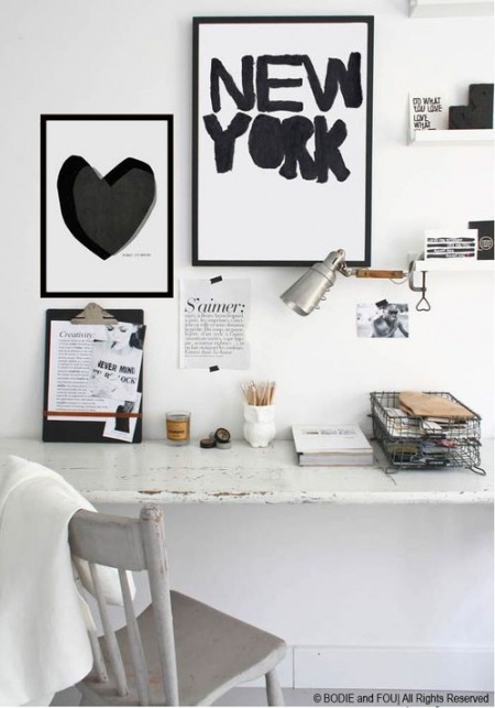 Białe biurko i białe krzesło postarzane w stylu schabby,druciany pojemnik na dokumenty,biało-czarne plakaty i typografie na białej ścianie nad biurkiem