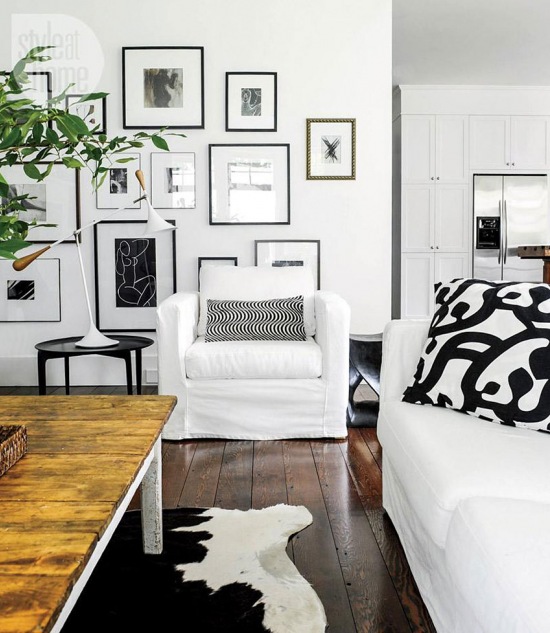 Galeria biało-czarnej fotogreafi i grafik na białej ścianie w salonie z białymi fotelami i sofą i rustykalnym stołem z drewnianym blatem
