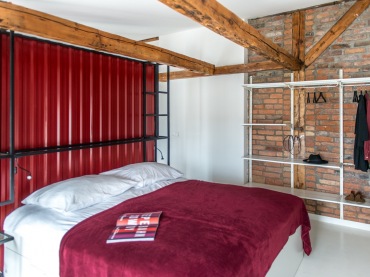 Na ścianie z czerwonymi cegłami w sypialni zorganizowano małą garderobę. Tworzą ją białe wieszaki i półki, które stylem...