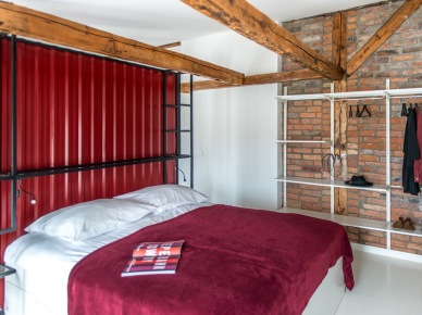 Na ścianie z czerwonymi cegłami w sypialni zorganizowano małą garderobę. Tworzą ją białe wieszaki i półki, które stylem przypominają klimat nowoczesnej lub industrialnej...