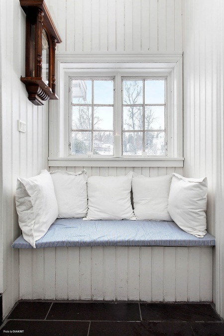 Pomysłowe  siedzisko z białych desek i poduszek pod małym oknem