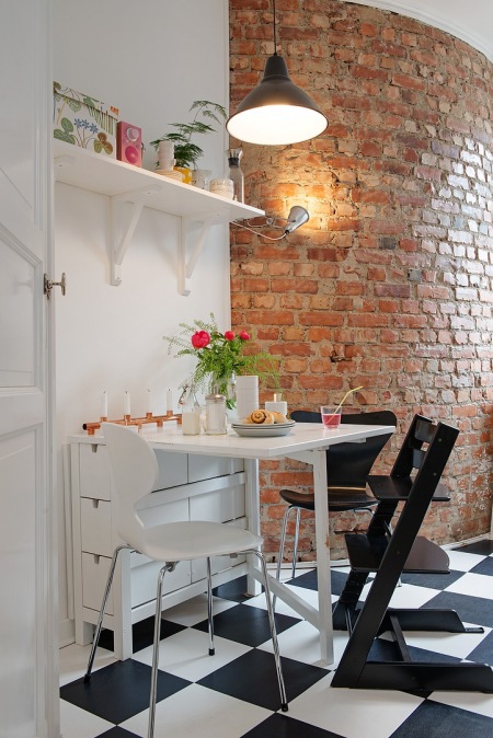Ściana z czerwonej cegły w kuchni,białe półki wiszące,rozkładany biały stół z szufladkami,białe iczarne krzesła Amadeo,czarne drewniane krzesło skandynawskie