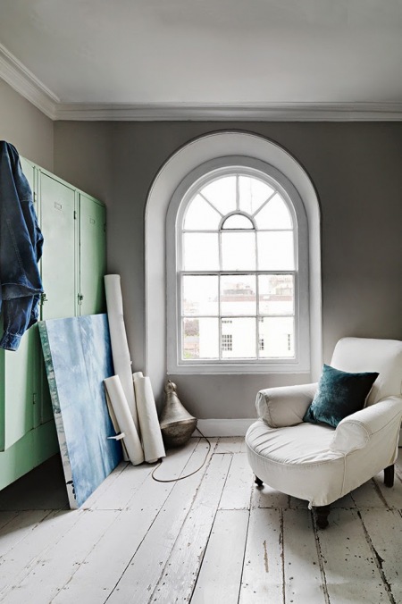 Szare ściany,turkusowa metalowa szafa,biały fotel na kółkach i okno z łukiem