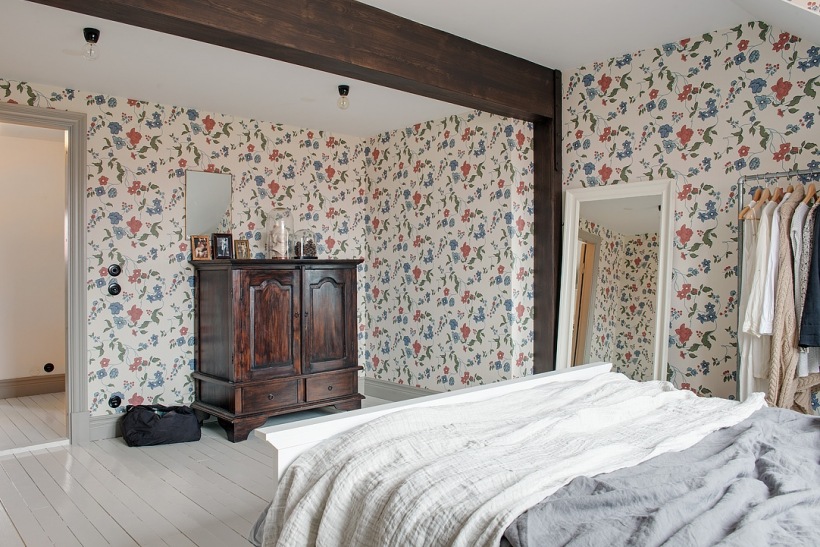 Drewniane ciemnobrązowe belki w sypialni,kwiatowa tapeta ścienna w niebiesko-czerwonym kolorze,retro komoda bieliźniarka przy ścianie,duże białe lustro stojące