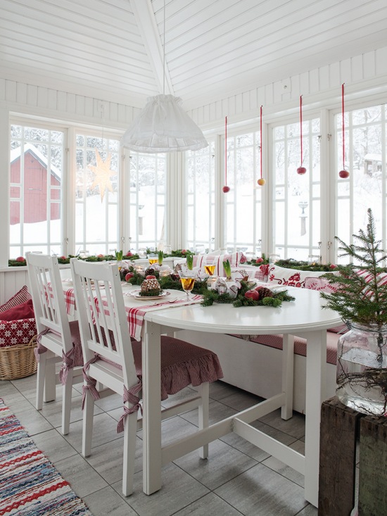 Biały drewniany stół z krzesłami w stylu skandynawskim,ławki białe ze świątecznymi poduszkami,biało-czerwone dekoracje świąteczne na wstążkach