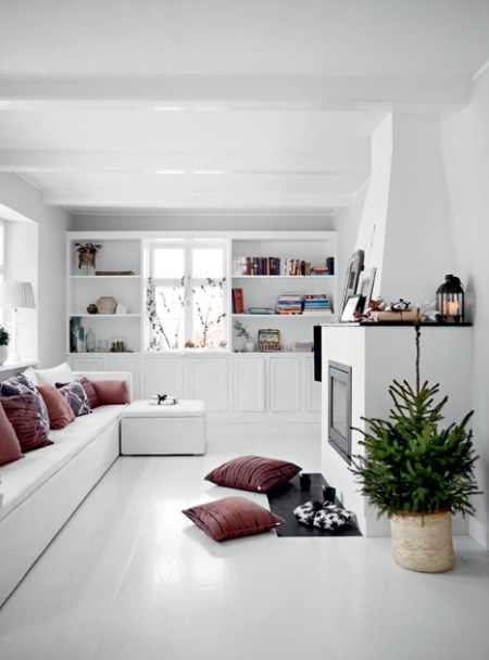 Biały salon jkominkowy z zabudowanymi siedziskami z poduchami i pufą,biała podłoga i półki wokół okna i malinowo-wrzosowe poduszki dekoracyjne