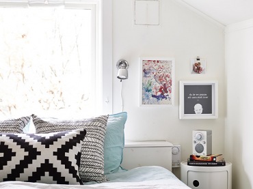 Owalna szafka nocna w kolorze białym, grafiki na ścinach i lampka zamontowana w ścianie to pomysł na urządzenie wąskiej przestrzeni w...
