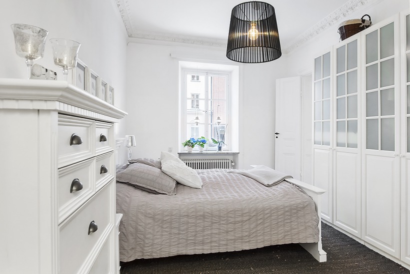 Biała komoda z szufladami,nowoczesna czarna lampa,szafa z witrynami i szara narzuta na łóżku w skandynawskiej sypialni