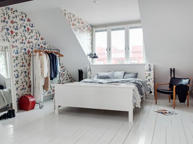Białe łóżko,stojący metalowy wieszak na kółkach,drewniane ciemnobrązowe belki w sypialni,kwiatowa tapeta ścienna w niebiesko-czerwonym kolorze, duże białe lustro stojące (25941)
