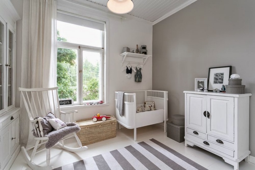 Biały fotel na płozach,biała komoda i łóżeczko w skandynawskiej aranżacji pokoju dla dziecka,szara śćiana i dywan w szaro-białe paski