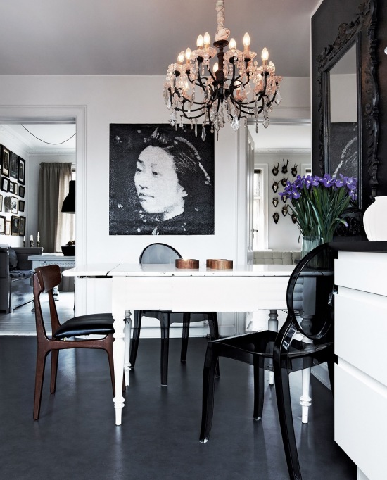 Stylowy żyrandol,biały rozkładany stół w tradycji skandynawskiej,czarne przeźroczyste krzesła z tworzywa,czarna podłoga z  żywicy i czarno-biała fotografia na ścianie