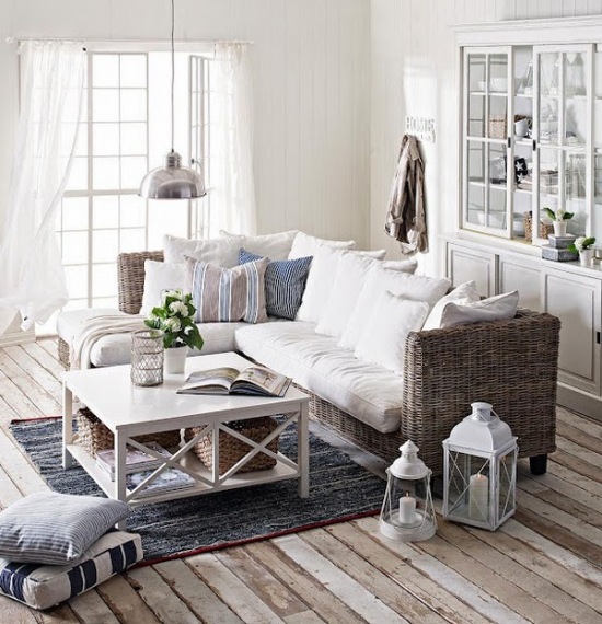 Prosta sofa kanapa z wikliny z białymi i szaro-niebieskimi poduszkami,białe lampiony i latarenki,biały kwadratowy stolik i biała witryna