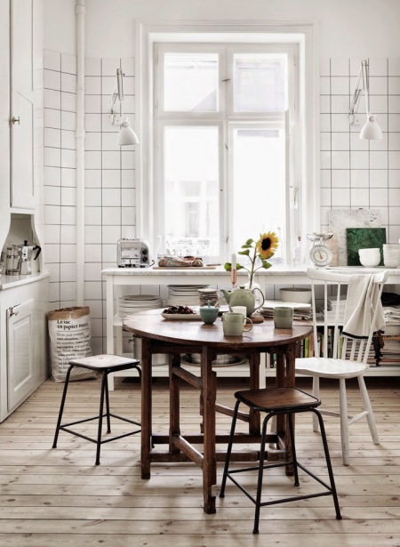 Podłoga z   desek z surowego drewna,białe płytki na ścianie,biała drewniana konsola z półkami,okragły drewniany stół rozkładany w kuchni