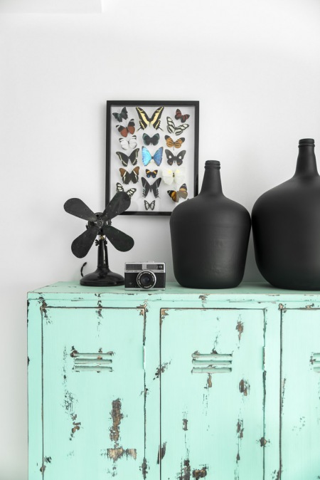 Turkusowa komoda z metalu,czarne pękate wazony i dekoracja na ścianę z kolekcją motyli