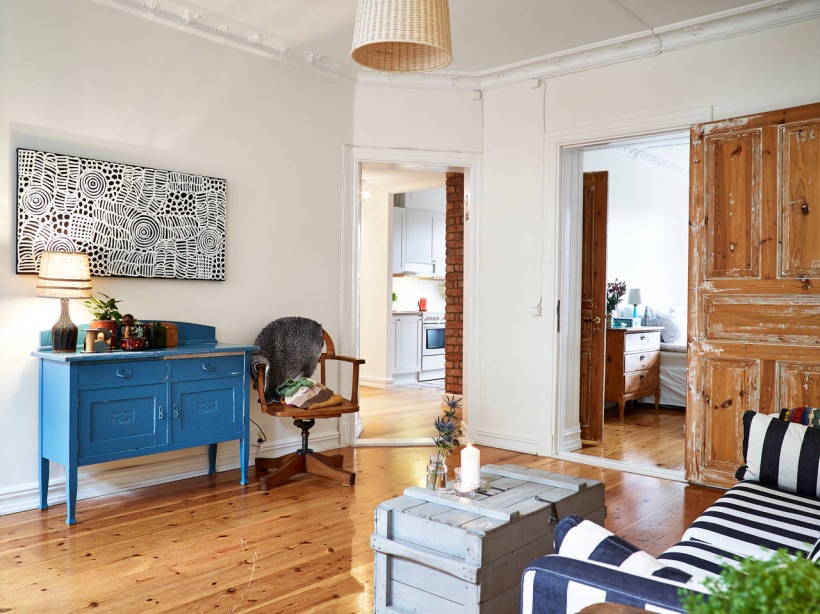 Drewniane bielone drzwi vintage,niebieska komoda,bambusowa lampa wisząca,szara skrzynia w roli stolika i pasiasta sofa w salonie
