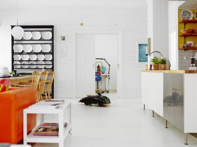 Serwantka z pólkami na białe talerze,biała podłoga, pomarańczowe sofy i biała kuchnia w otwartej zabudowie wnętrza (24658)