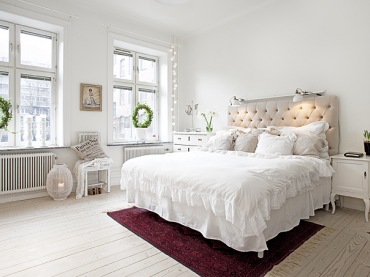 Jasna, niemal zupełnie biała sypialnia z wielkim łożem z pikowanym wezgłowiem.Mocniejsze kolory wprowadzają tu bordowy dywan pod łóżkiem i zielone kwiaty ułożone w fantazyjne...