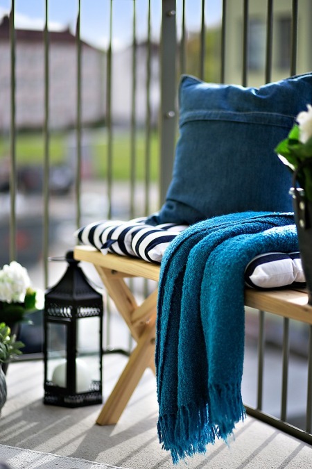 Czarny lampion,drewniany fotel i turkusowo-niebieskie poduszki i pledy na balkonie