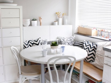 Jasna jadalnia z białym okrąglym stołem i białymi krzesłami. Dekorację stanowią tu poduszki w czarno – białe,...