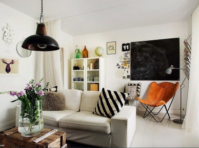 Jasny salon w skandynawskim stylu z szara sofę został skontrastowany przez czarna lampę i tablicę. Poduszka w biało-czarne paski to fajny akcent, nie wspominając o rdzawym fotelu...