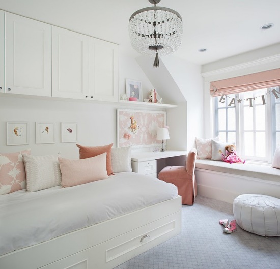 Białe łóżko z szafkami na ścianie,małe biurko i zabudowa z siedziskami przy oknie z różową roletą i poduszkami dekoracyjnymi