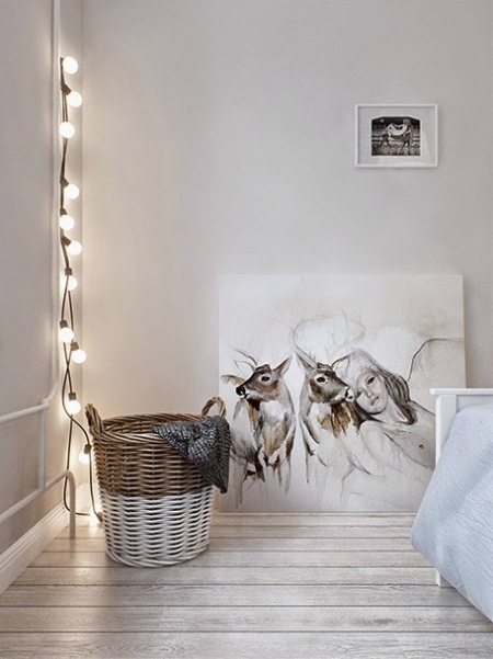Bielone deski na podłodze,girlanda z żarowkami na kablu,skandynawskie ilustracje i biało-beżowy kosz wiklinowy z uszami w sypialni