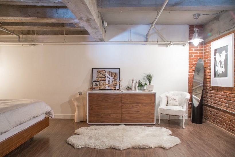 Biały dywan futrzak,białe ściany,przemysłowy sufit z betonowymi belkami,nowoczesna biało-drewniana komoda,stylowy biały fotelik i ściana z czerwonej cegły w sypialni