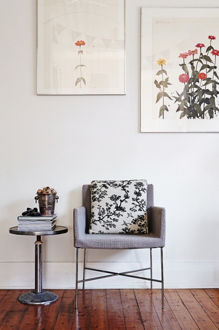 Nowoczesny szary fotel na metalowych nogach, okragły stolik pomocniczy i kolorowe artprinty na białej ścianie