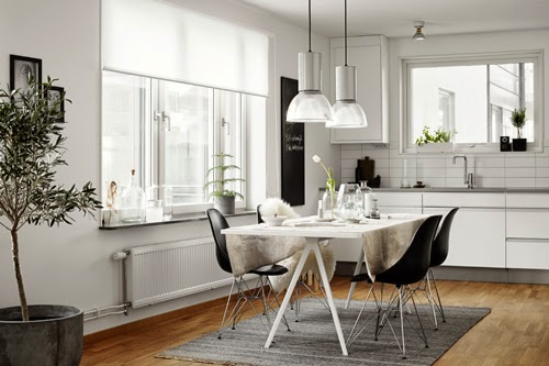 Biały skandynawski stół na kozłach,czarne nowoczesne krzesła na metalowych krzyżakach,szary tkany dywan,nowoczesne szklano-metalowe lampy nad stołem w białej kuchni