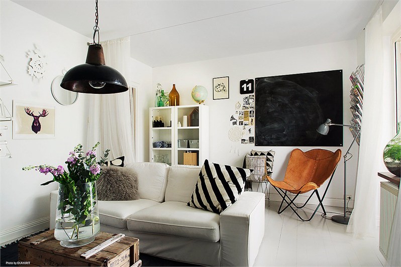 Mały bialy salon z czarną tablicą, fotelem butterfly,lampą industrialną i drewnianą skrzynią