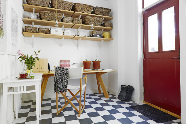 Czerwone drzwi,drewniane półki z wiklinowymi koszykami,drewniane biurko i podłoga w biało-czarna szachownicę,