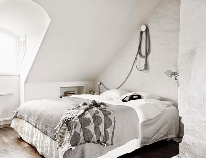Biało-szara narzuta na łóżku w skandynawskiej sypialni z ciekawym mocowaniem żarówki na kablu na ścianie nad łóżkiem