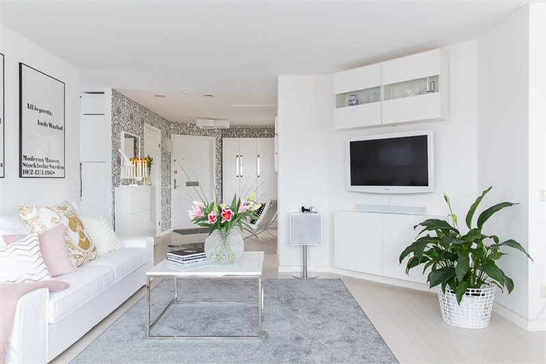 Szary dywan,kwadratowy stolik na chromowanych nogach,biała sofa z różowo-pudrowymi poduszkami w otwartym salonie