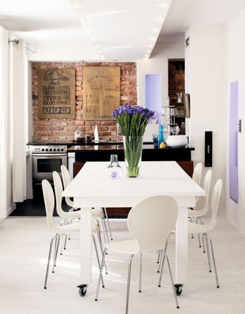 Bialy stół na kółkach,nowoczesne krzesła,,czerwona cegła na ścianie i stalowe szafki kuchenne w aranżacji nowoczesnej kuchni