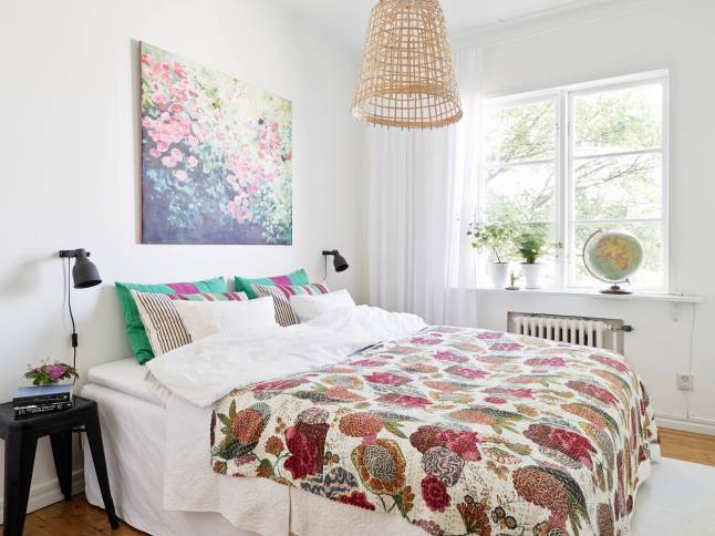 Pleciony abażur wiszącej lampy,narzuta i obraz z motywem kolorowych kwiatów,turkusowo różowe poduszki w sypialni