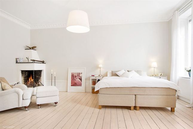 Beżowe tapicerowane łóżko,podłoga z naturalnych desek,fotel z podnóżkiem na kółkach w sypialni z murowanym kominkiem