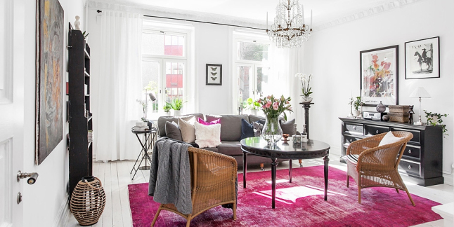 Eklekryczny salon z różowym dywanem i czarnym okrągłym stołem, wiklinowymi fotelami i skandynawskim lampionem