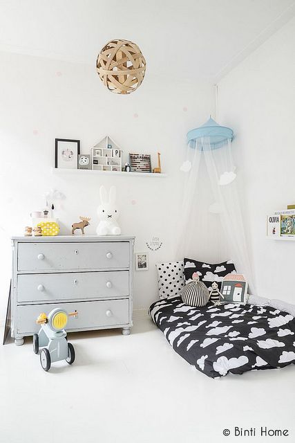 Jasnoszara komoda z szufladami w białym pokoju dziecięcym,biało-czarna pościelk w chmurki i wąska półeczka na ścianie