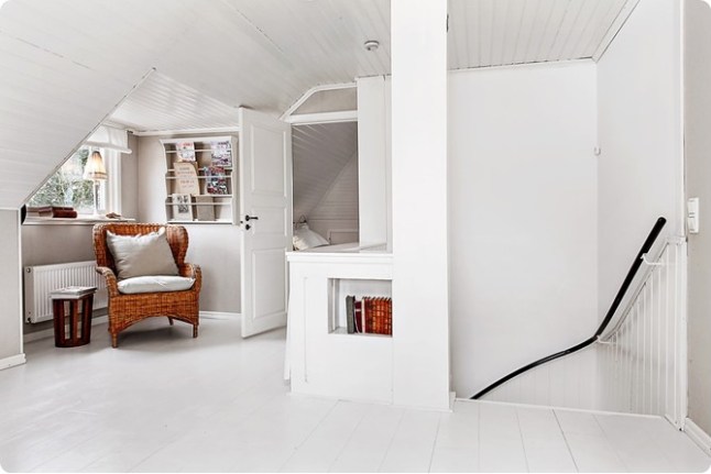 Rattanowy duży fotel z poduszkami,półka na czasopisma na ścianie na białym poddaszu w skandynawskim domku