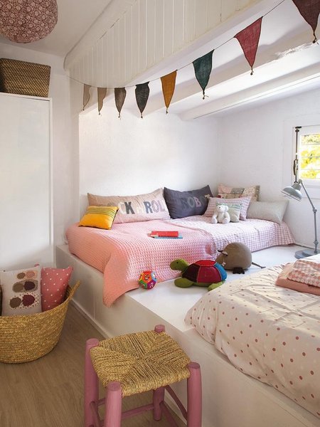 Dziecięce łóżka na białym podeście,kolorowe proporczyki ,różowy taboret z sizalowym siedziskiem,kosz z liści bambusa,kolorowe poduszki w pokoju dla dziecka