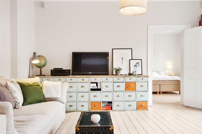 Turkusowo-miodowe szuflady w komodzie,skandynawskie grafiki,granatowy kufer stolik i biała sofa z zielonymi poduszkami